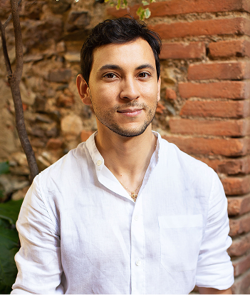 Portrait de Noham portant une chemise blanche, assis devant un mur en briques avec un léger sourire.